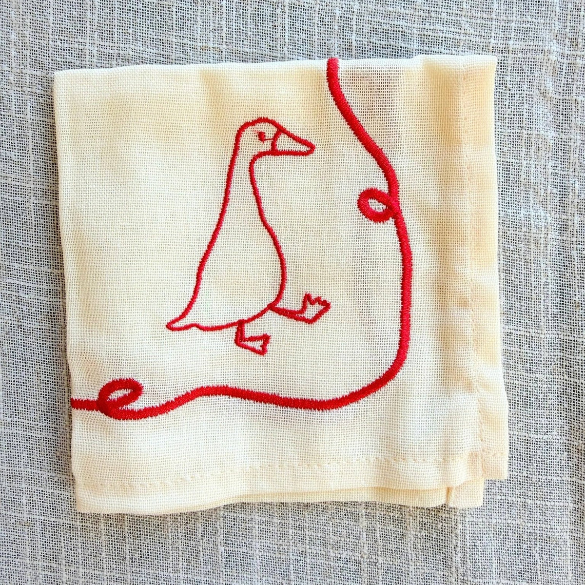A folded cream-coloured linen napkin featuring a red embroidered goose and a red looped line design, displayed on a textured gray linen background.