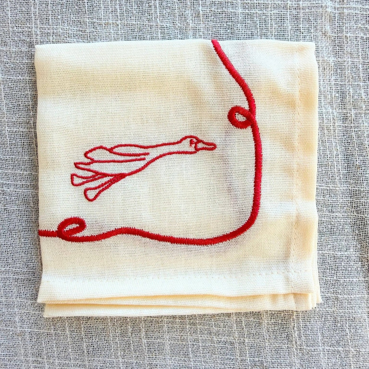 A folded cream-colored cocktail napkin with red embroidered duck, displayed on a textured gray linen background.