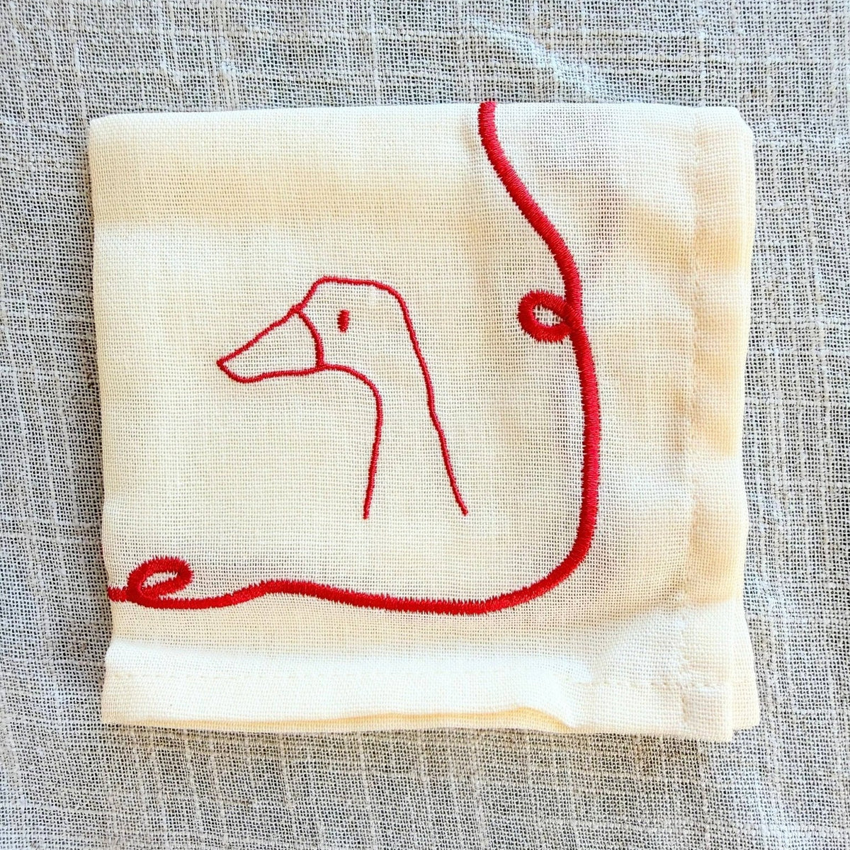 A folded cream-coloured cocktail napkin featuring a minimalist red embroidered line drawing of a goose, placed on a textured gray linen surface.