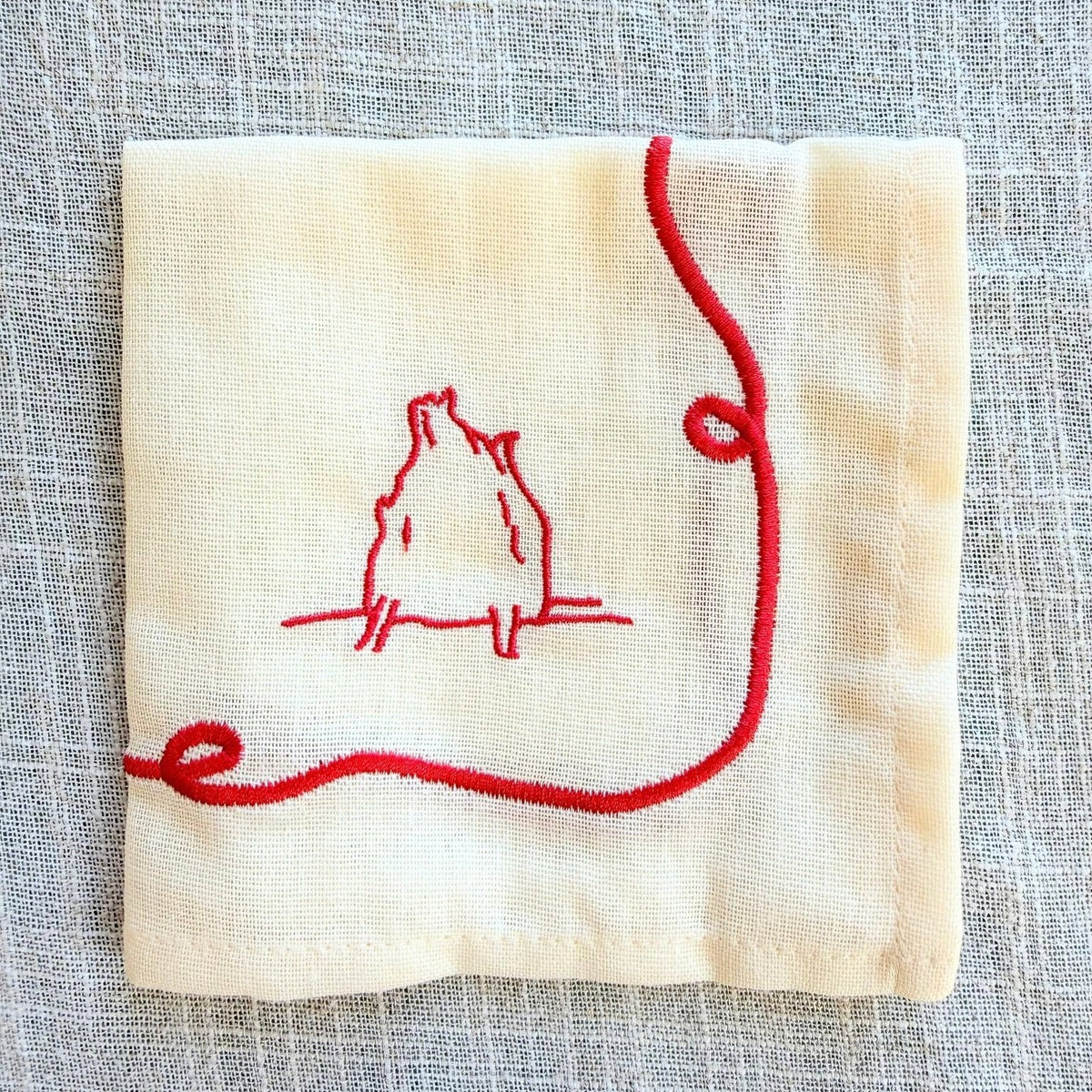 A square cream-colored linen napkin featuring a red embroidered duck design  with a red loop extending from the bottom left, laid flat on a textured gray linen background.