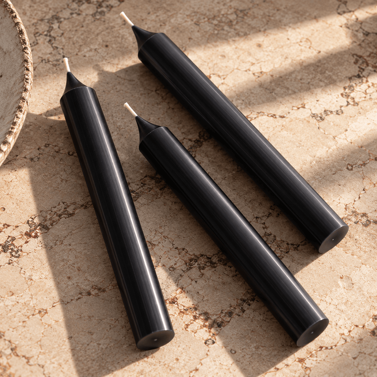 Three black taper candles with white wicks arranged diagonally on a textured beige stone surface with soft shadows.