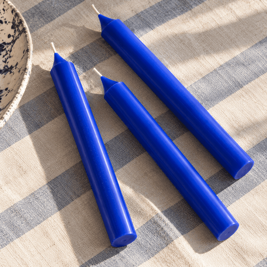 Three cobalt blue taper candles arranged diagonally on a beige and blue striped fabric surface.