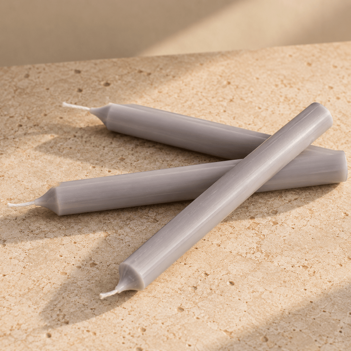 Three pale grey taper candles arranged in an X pattern on a textured beige stone surface with soft shadows.