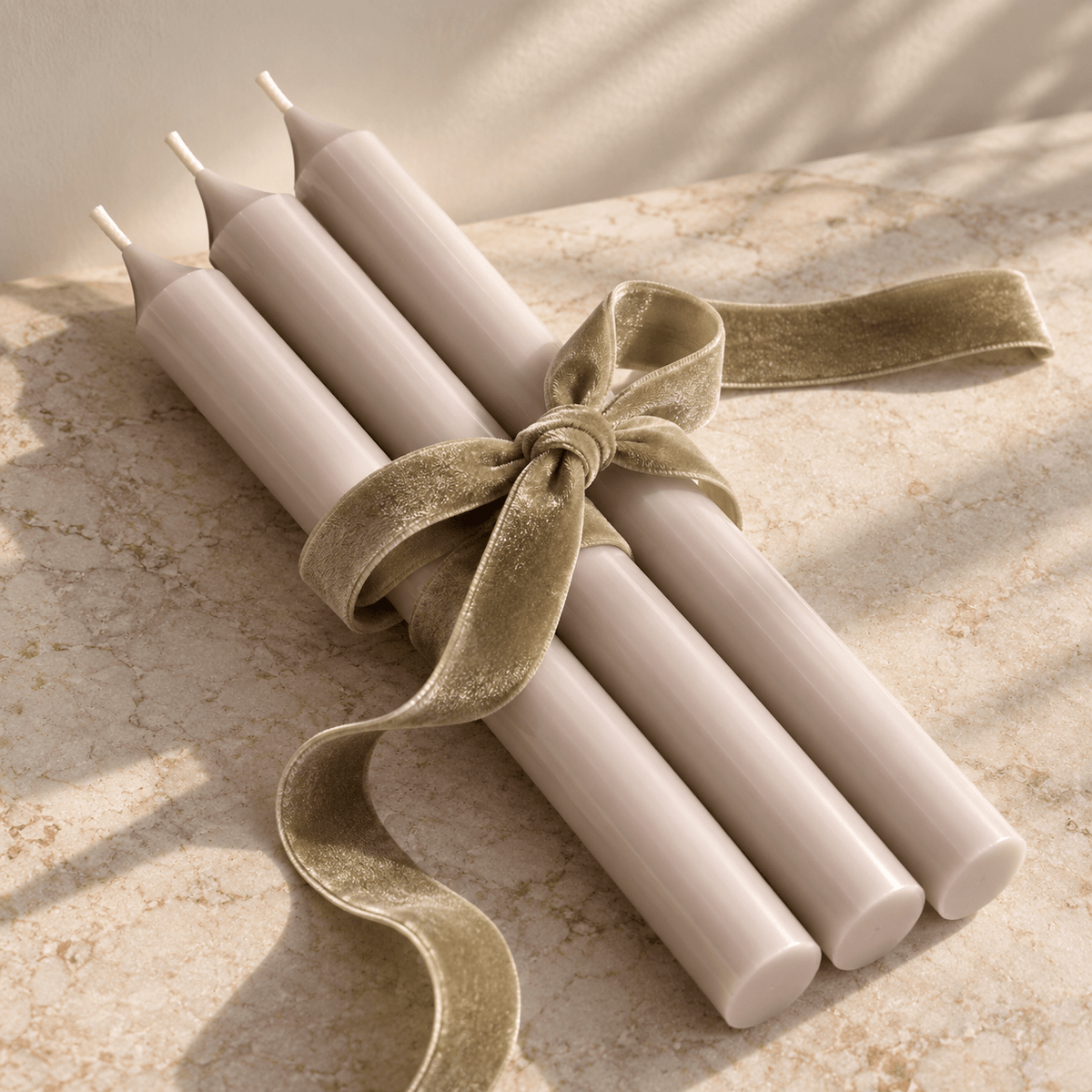 Four tapered pale grey candles tied together with a green-coloured velvet ribbon bow, arranged on a light stone surface with soft shadows.