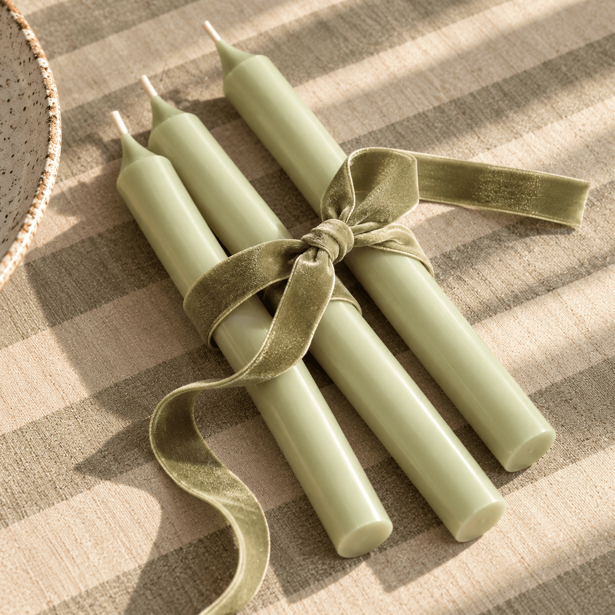 Four pale green taper candles tied together with a matching sage green velvet ribbon, arranged on a beige and taupe striped fabric surface.
