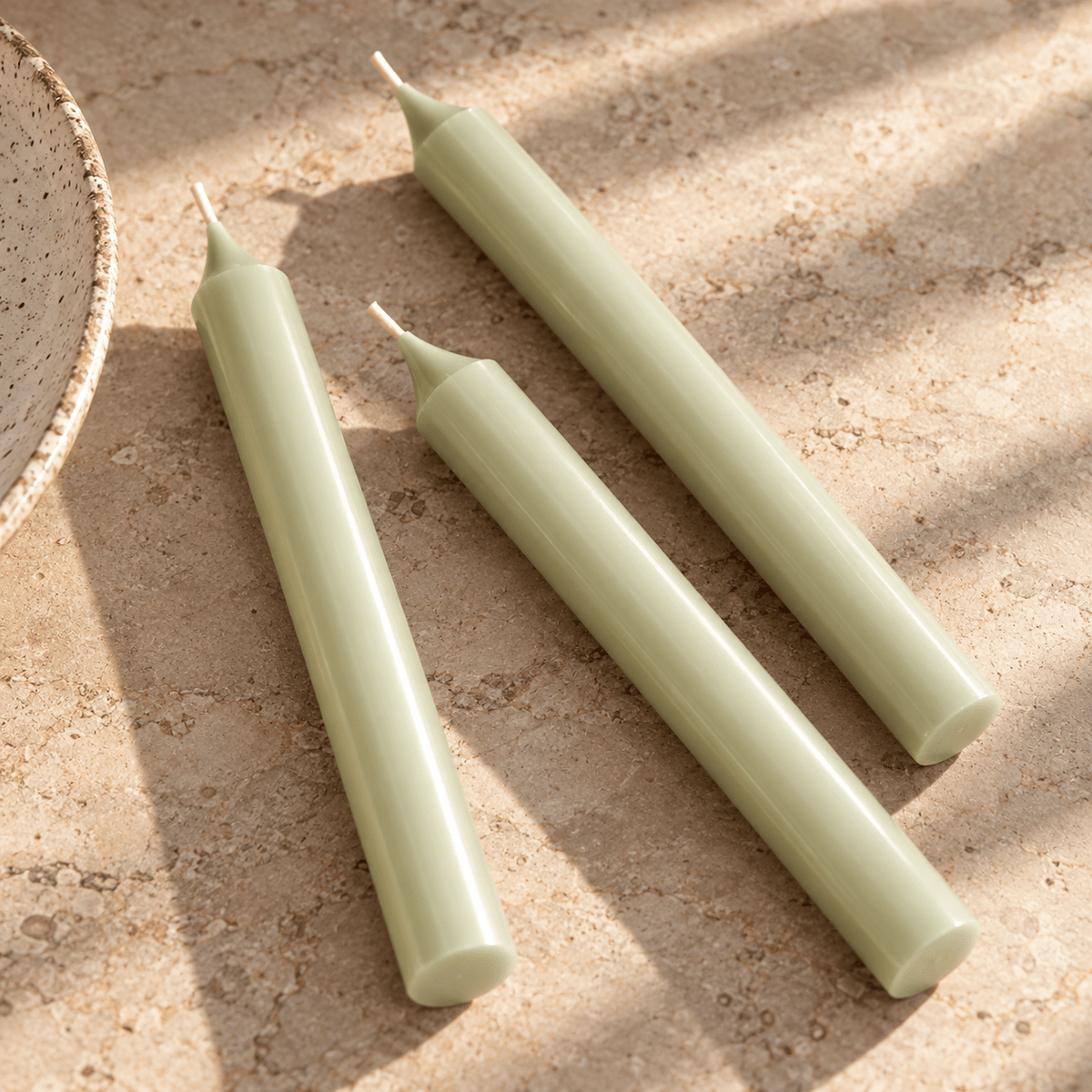 Three pale green taper candles arranged diagonally on a textured beige stone surface with soft shadows.