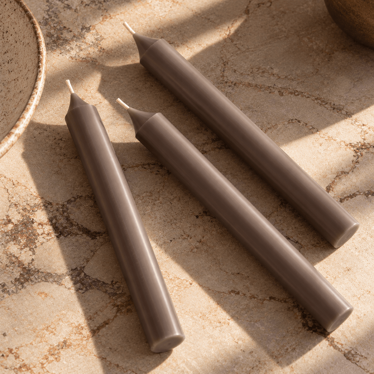 Three tapered candles in a warm taupe-brown colour arranged diagonally on a textured beige stone surface with natural shadows.