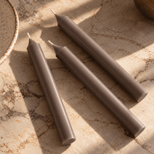 Three tapered candles in a warm taupe-brown colour arranged diagonally on a textured beige stone surface with natural shadows.