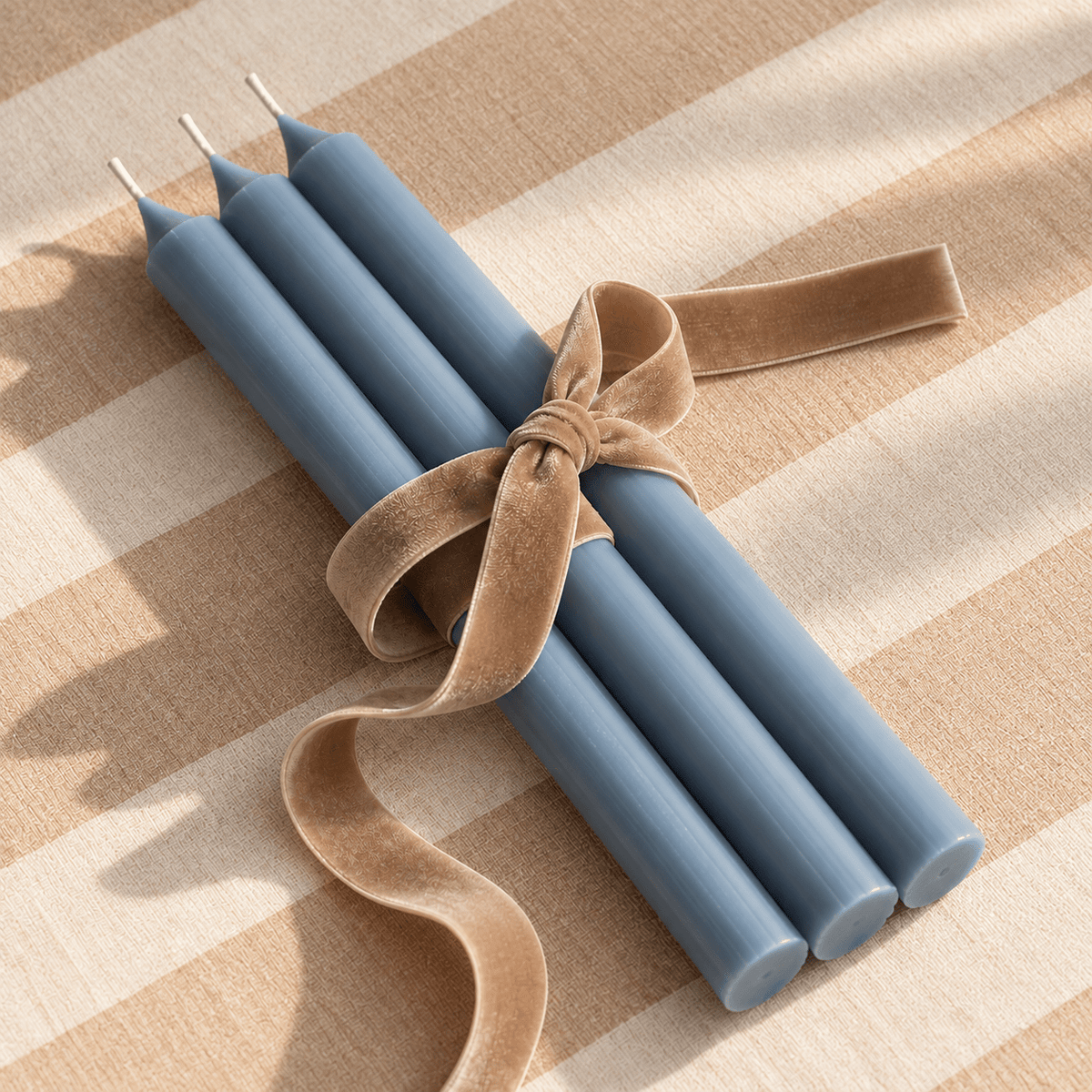 Four blue taper candles tied together with a tan velvet ribbon, placed on a beige and cream striped fabric surface.