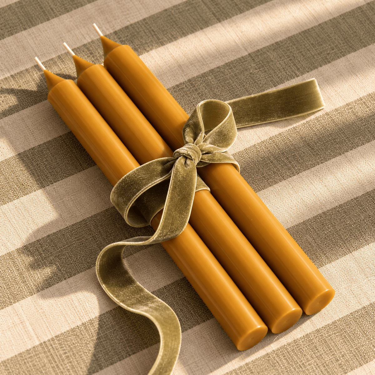 Four yellow ochre taper candles tied together with a wide olive-green velvet ribbon, arranged on a beige and olive striped fabric background.