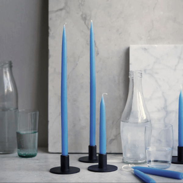 Three tall blue taper candles in black metal holders arranged on a light stone surface, with additional blue candles and clear glass bottles and glasses in a styled interior setting.