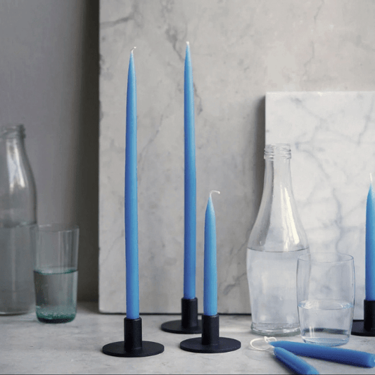Three tall blue taper candles in black metal holders arranged on a light stone surface, with additional blue candles and clear glass bottles and glasses in a styled interior setting.