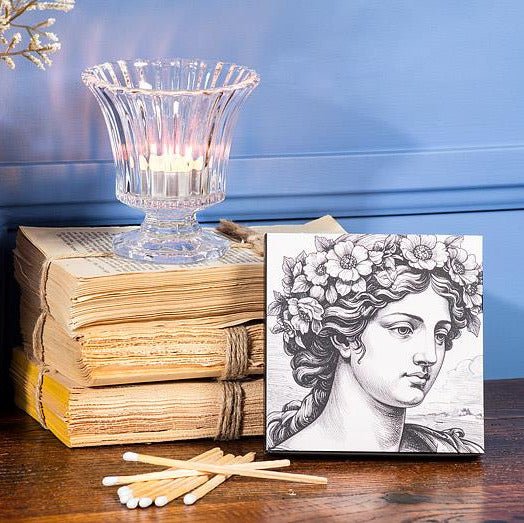 A square matchbox with a classical bust illustration, styled on a wooden table next to a stack of old books, a lit glass candle holder, and scattered matches against a blue paneled wall.