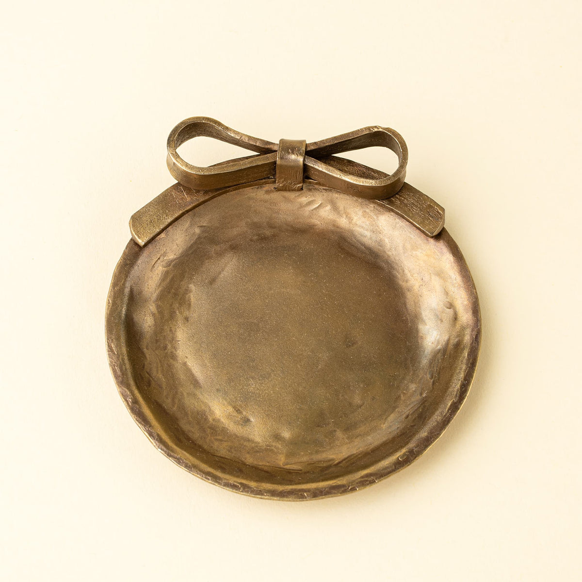 A round hammered metal dish with a bow detail on top, shown against a plain cream background.