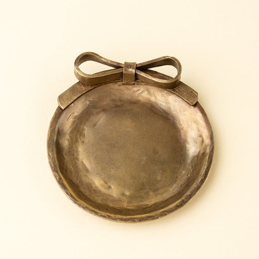 A round hammered metal dish with a bow detail on top, shown against a plain cream background.