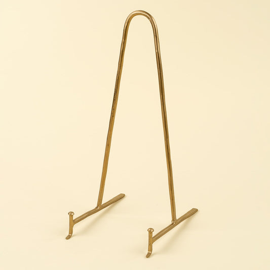 A gold-finished metal display stand with a curved top bar and two angled feet, shown on a plain warm cream background.