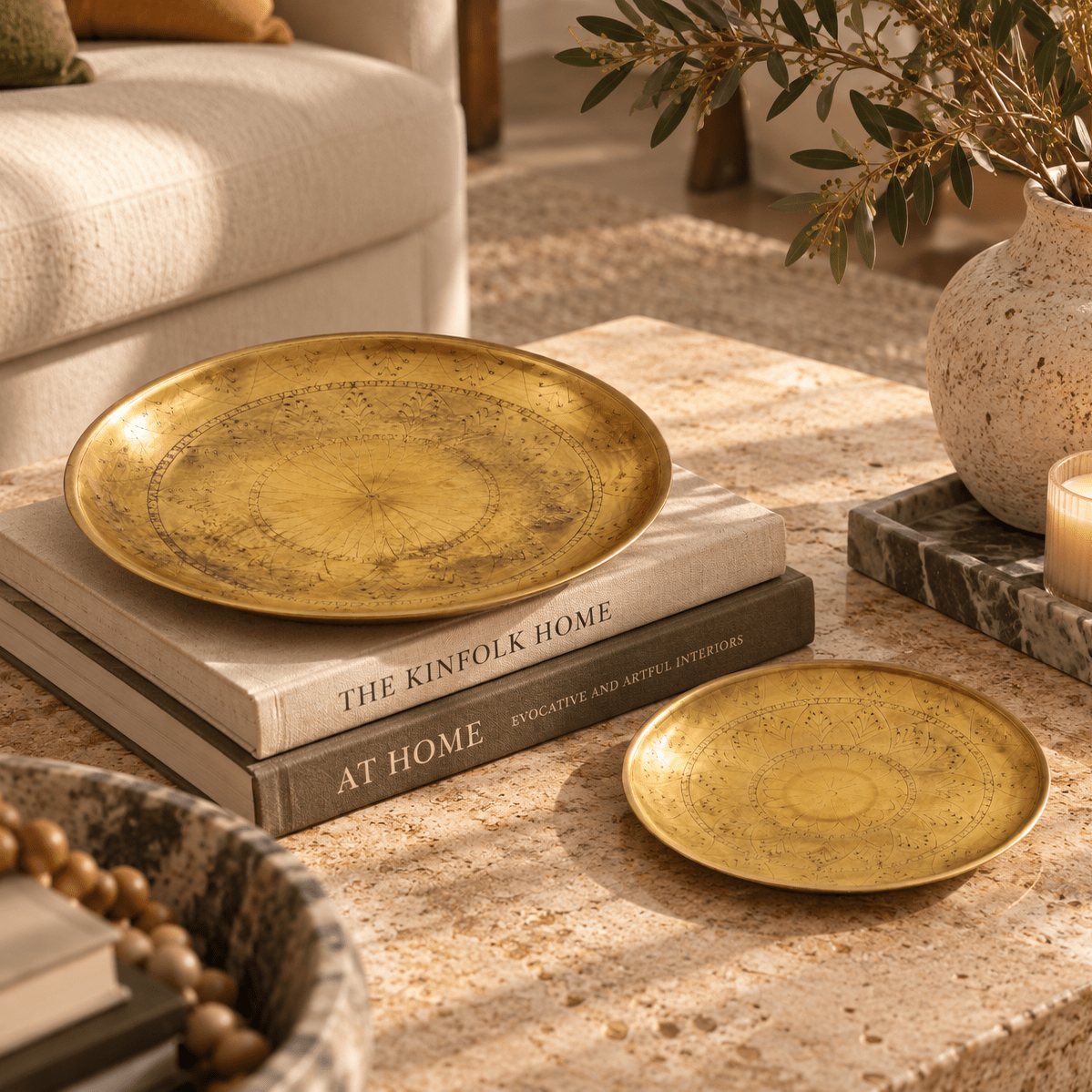 Two round brass decorative plates with a textured, speckled gold finish are styled in a warm, sunlit living room setting. One larger plate sits on a stack of books, and a smaller matching plate rests beside it on a stone surface.