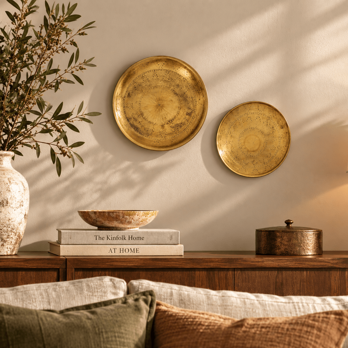 Two round distressed brass wall plates hanging on a beige wall in a styled living room setting with a wooden console table, decorative books, a vase with branches, and cushions in the foreground.