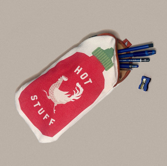 A fabric pencil pouch designed like a red hot sauce bottle with a white rooster graphic and the words 'HOT STUFF'. It is shown on a neutral beige background with blue pens and a blue sharpener peeking out from the top.
