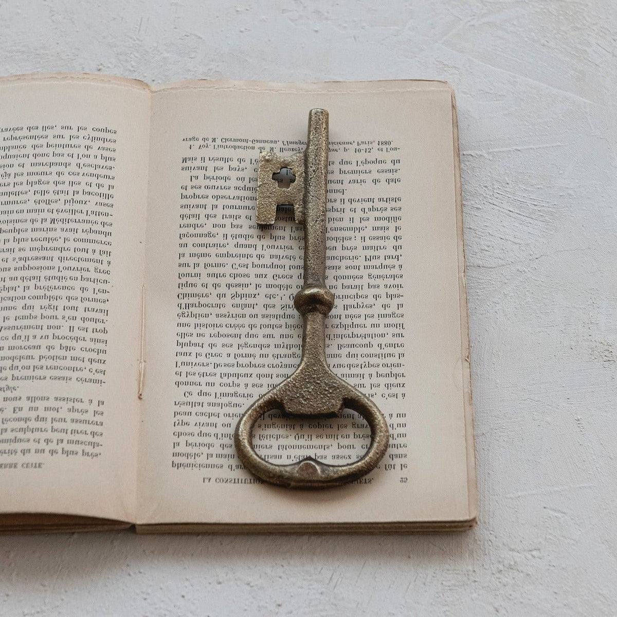 An antique-style metal key-shaped bottle opener, placed on top of an open vintage book with printed text, set against a light textured background.