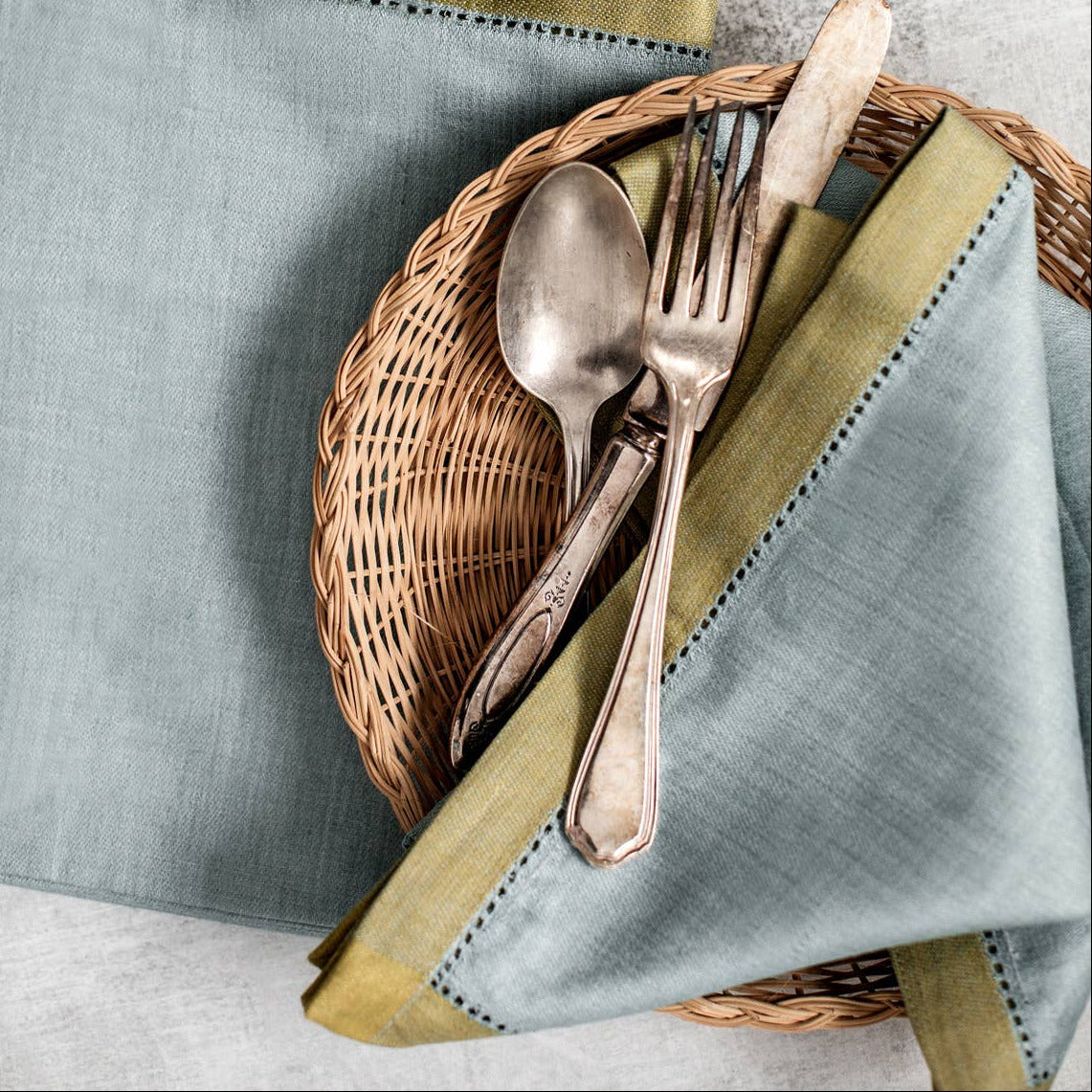Wicker basket with silverware on a light blue napkin, next to a ceramic vase on a textured surface.