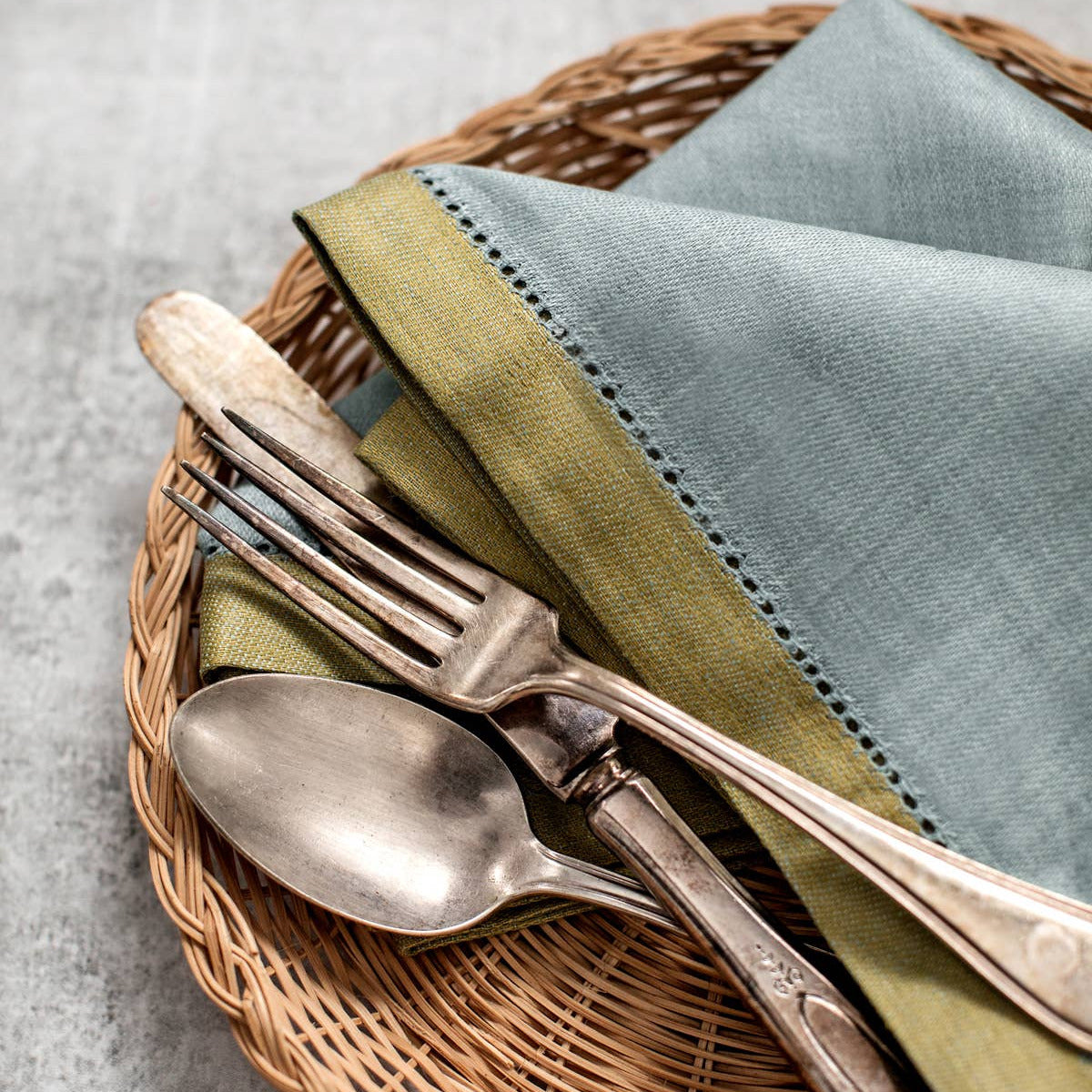 A styled place setting featuring a folded pale blue linen napkin with green stitching, set on a round woven wicker placemat with vintage silver-toned flatware.