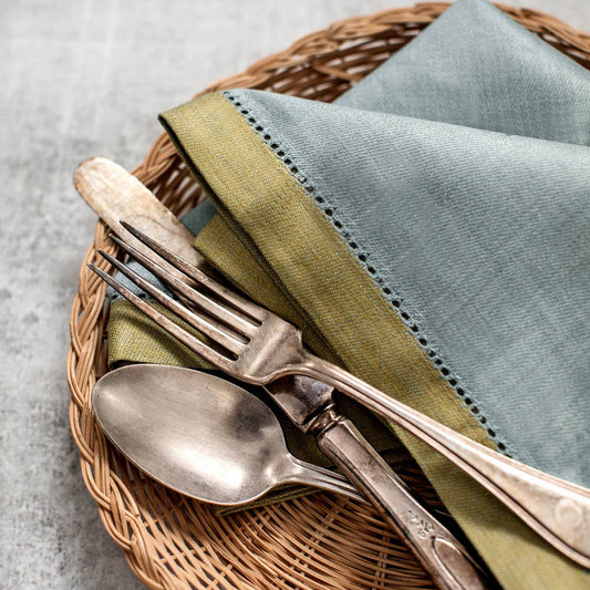 A styled place setting featuring a folded pale blue linen napkin with green stitching, set on a round woven wicker placemat with vintage silver-toned flatware.