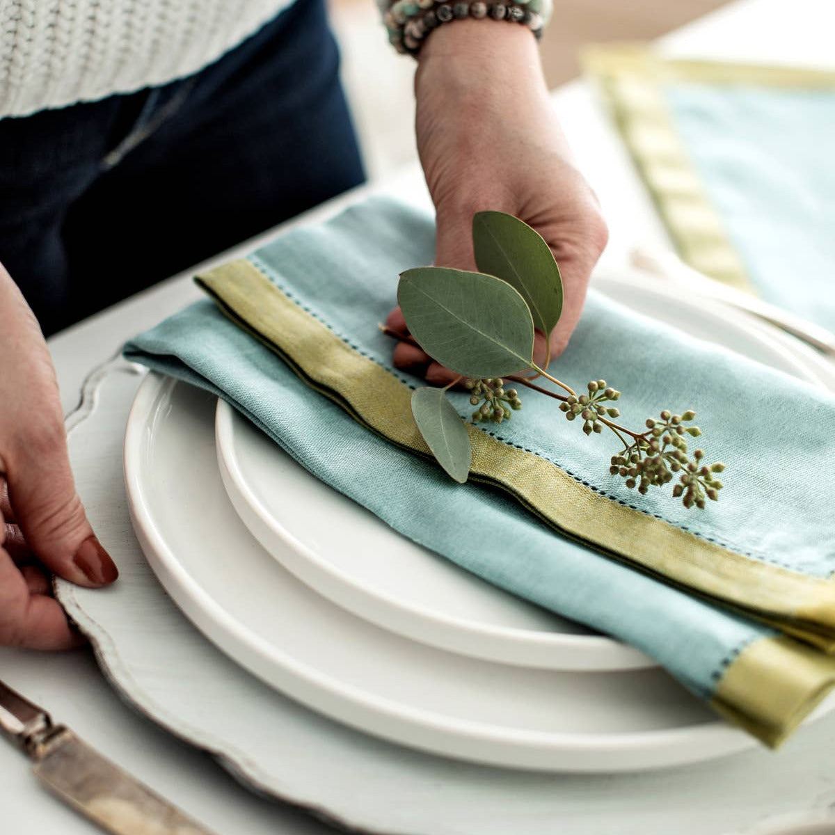 A close-up lifestyle shot of a table setting featuring a light blue linen napkin with a green hemstitch border, styled with white plates and a green eucalyptus sprig. A person's hands and torso are visible adjusting the napkin.