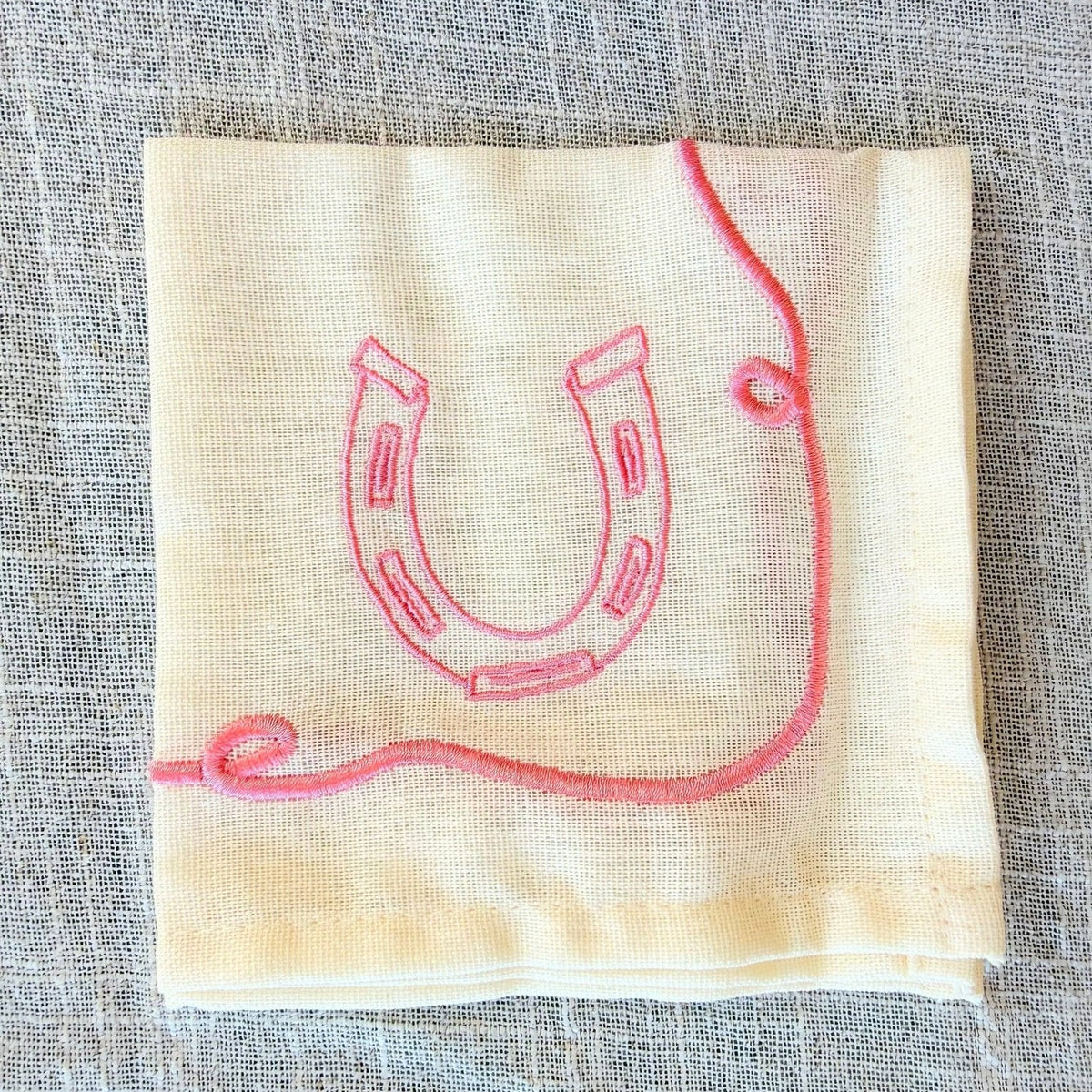 A folded cream-colored linen napkin with a pink embroidered horseshoe and loop design, displayed on a textured gray linen surface.