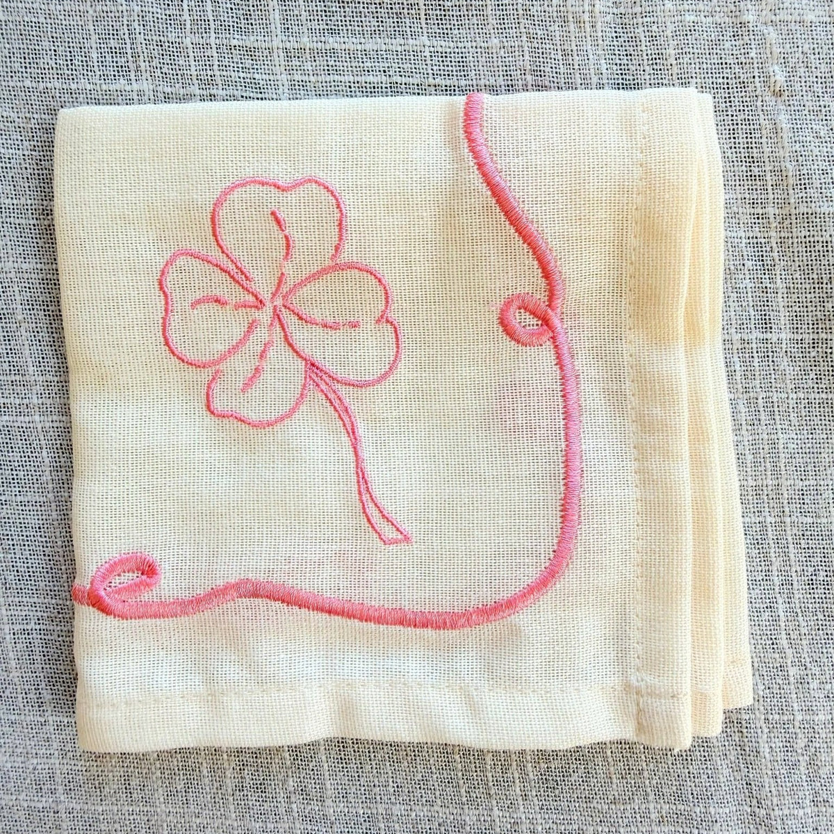 A folded cream-colored linen napkin featuring a pink embroidered four-leaf clover and a matching pink border design, displayed on a textured gray linen background.