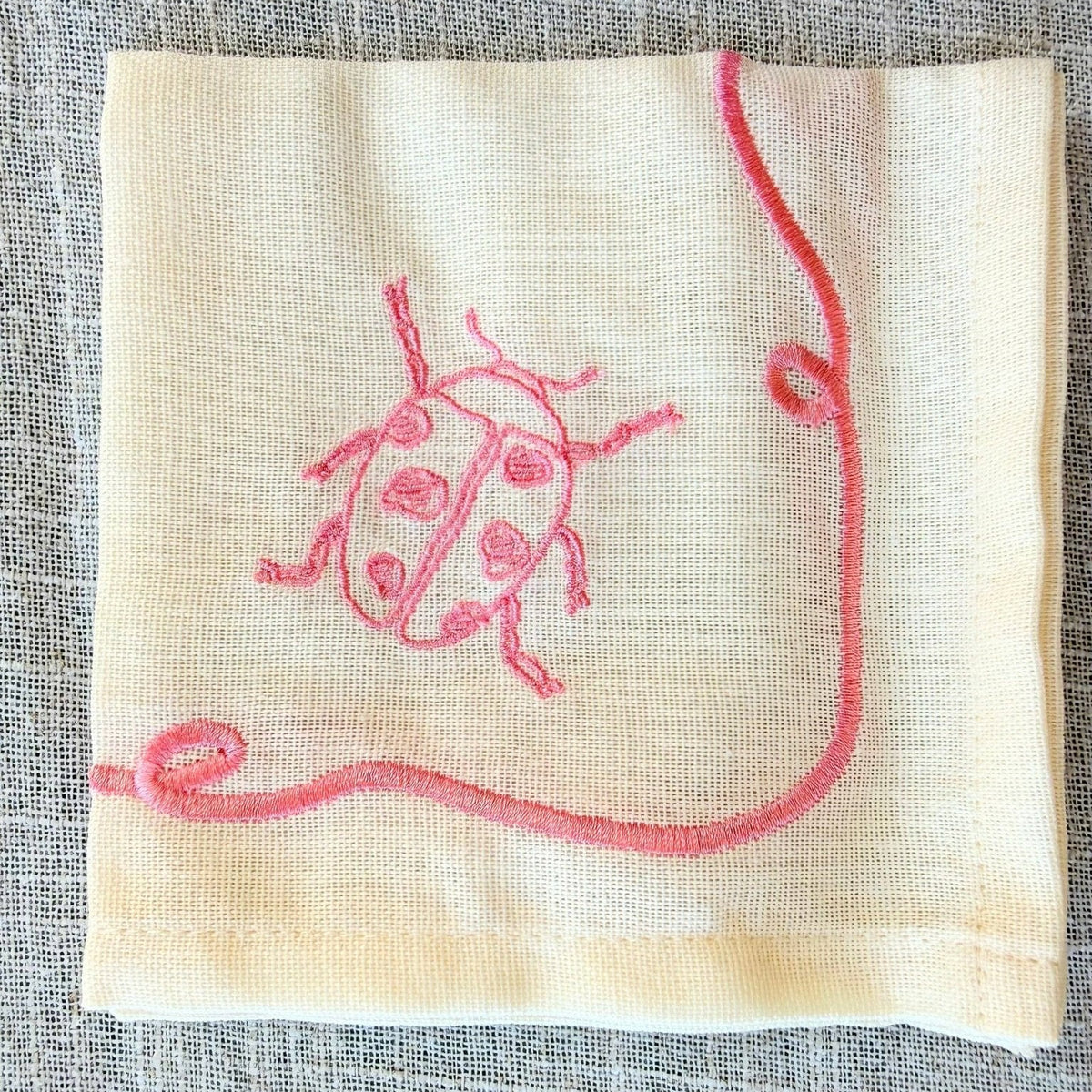 A square cream-colored cocktail napkin featuring a pink embroidered ladybug and a curved vine design, laid flat on a textured gray linen background.