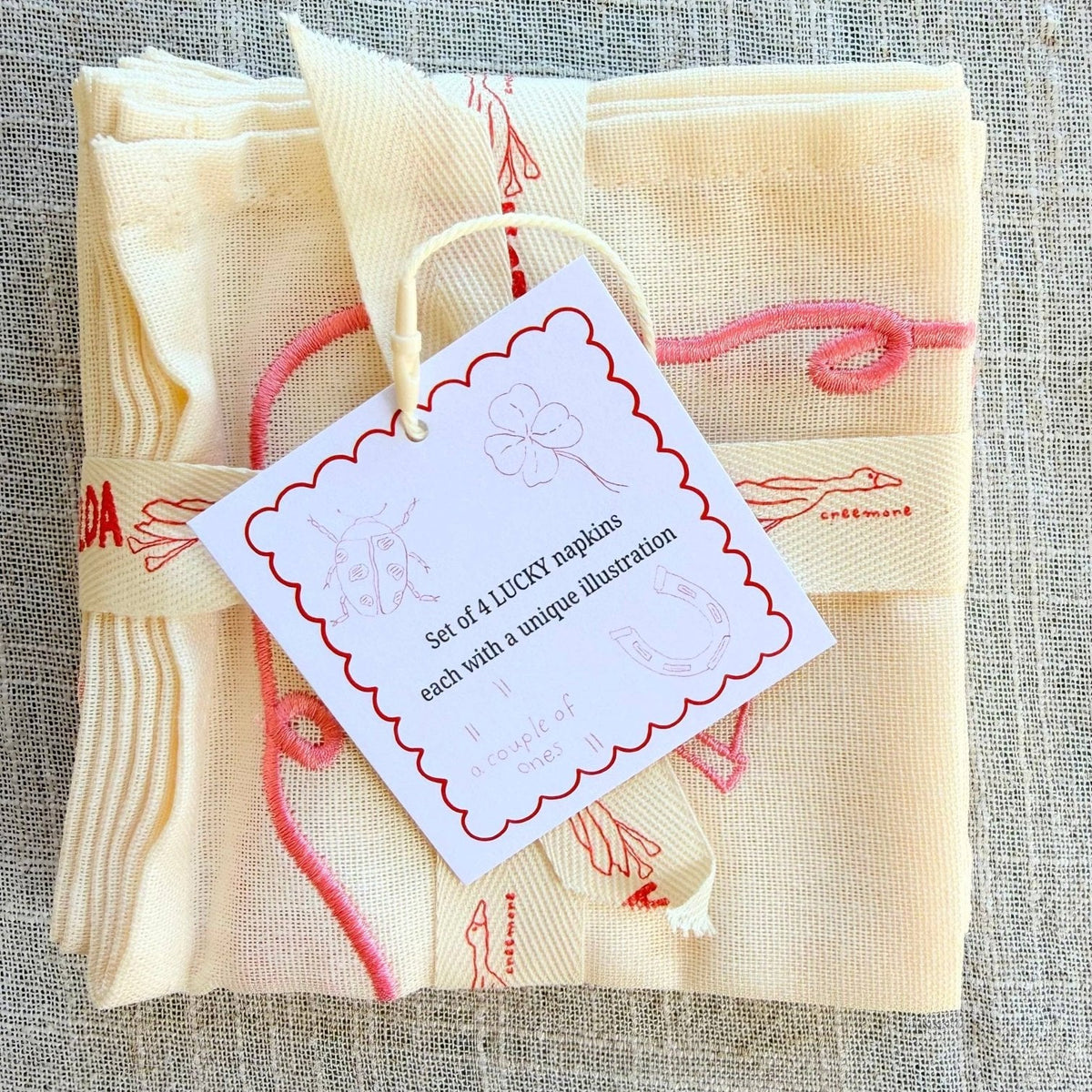 A folded set of four cream-colored cocktail napkins with pink embroidered good luck symbols, tied with a matching ribbon and a white paper tag.