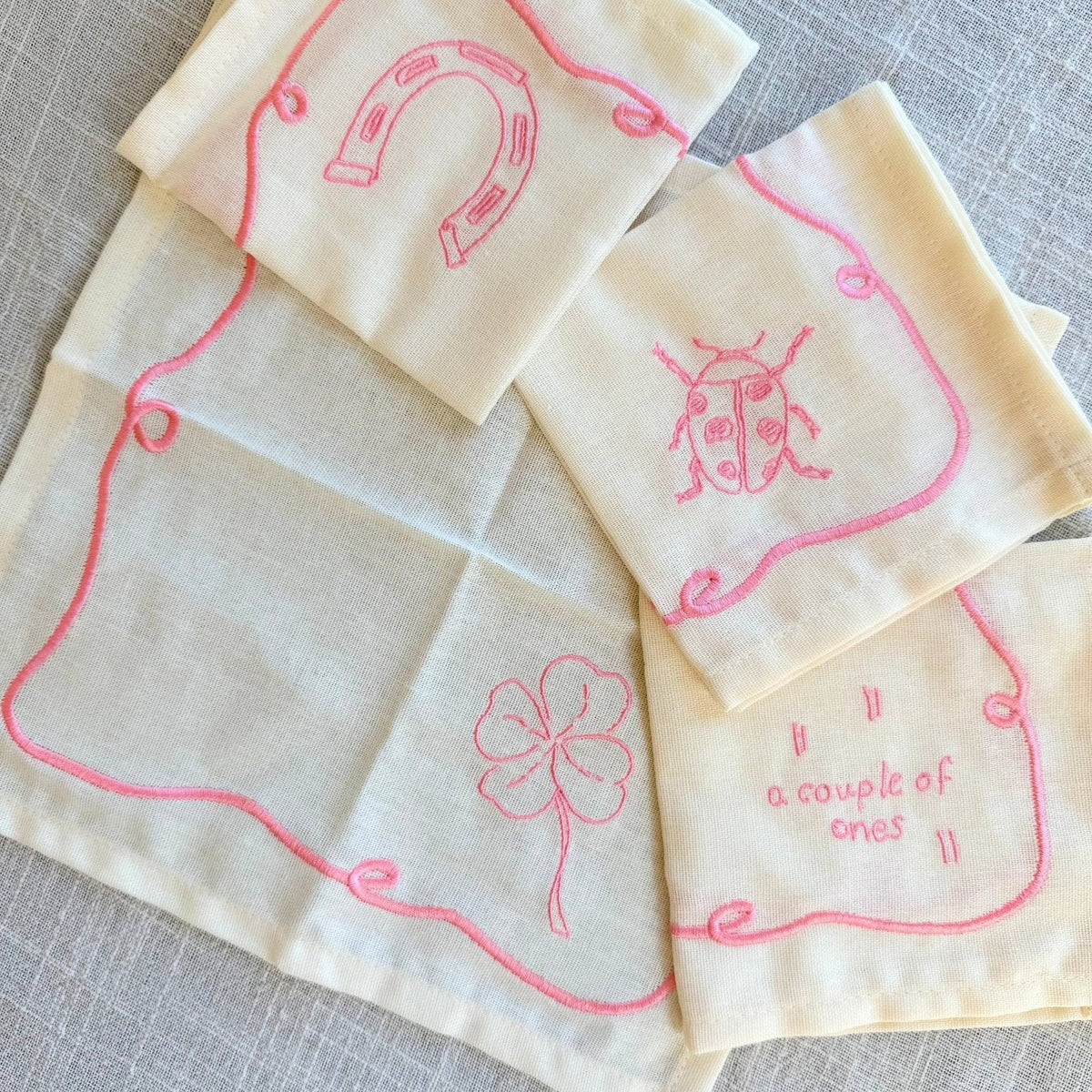 Four cream-colored cocktail napkins with pink embroidered designs, including a horseshoe, a ladybug, a four-leaf clover, and the phrase 'a couple of ones', arranged on a textured gray fabric background.