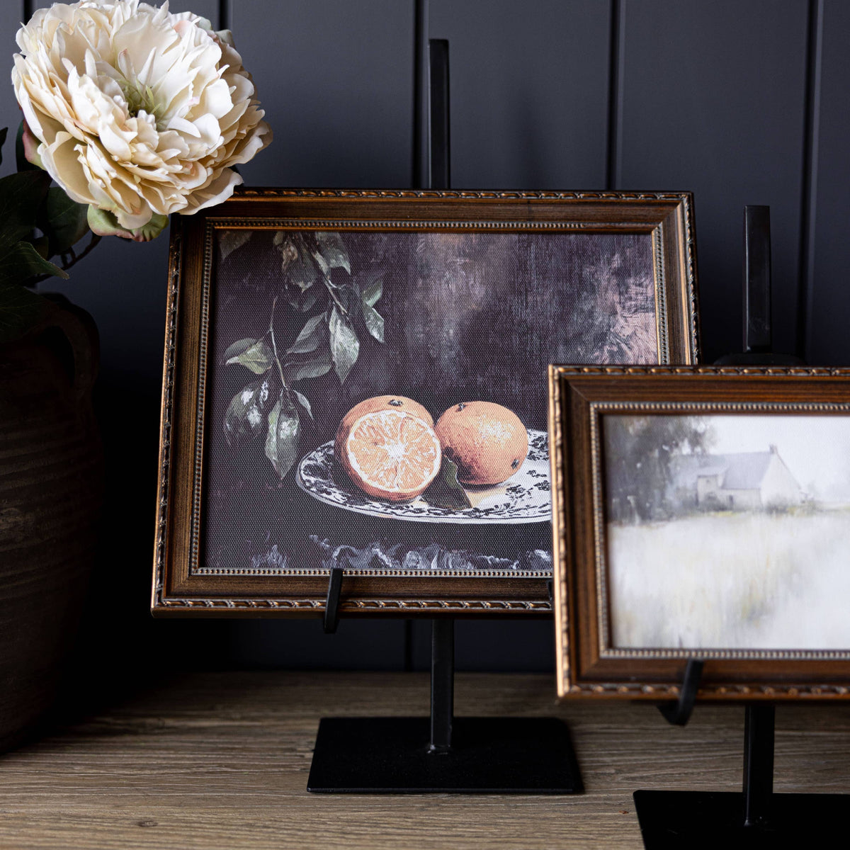Two framed artworks on art easel against a dark panelled wall.