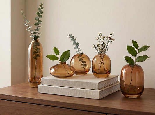 Four amber glass vases of different shapes and sizes arranged on a wooden surface with two stacked books. Three vases contain green eucalyptus stems, and one vase on the right holds a green leafy branch.