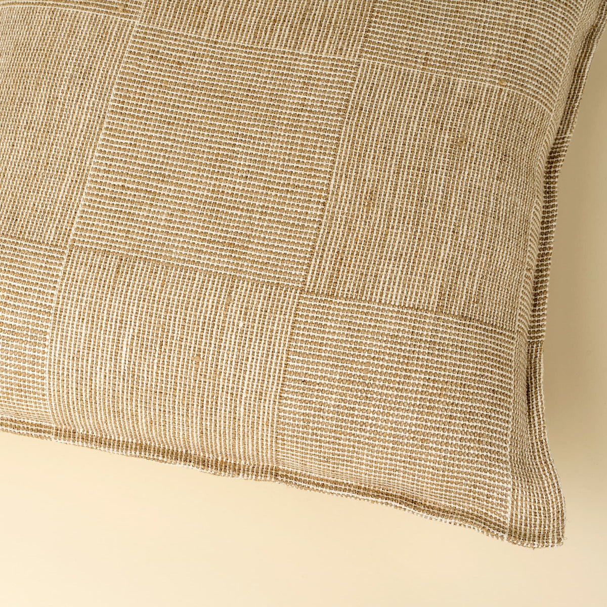 A close-up of a square throw pillow featuring a textured, woven patchwork design in cream and beige tones, set against a solid cream background.