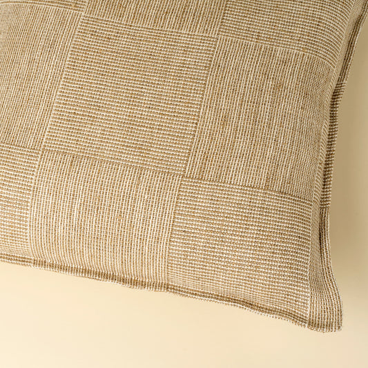 A close-up of a square throw pillow featuring a textured, woven patchwork design in cream and beige tones, set against a solid cream background.