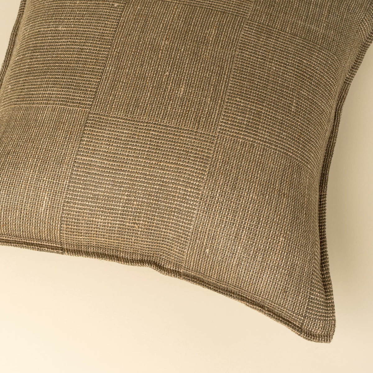 A close-up of a square throw pillow featuring a textured, woven patchwork design in olive and beige tones, set against a solid cream background.