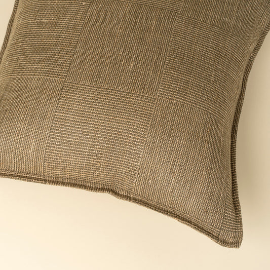 A close-up of a square throw pillow featuring a textured, woven patchwork design in olive and beige tones, set against a solid cream background.