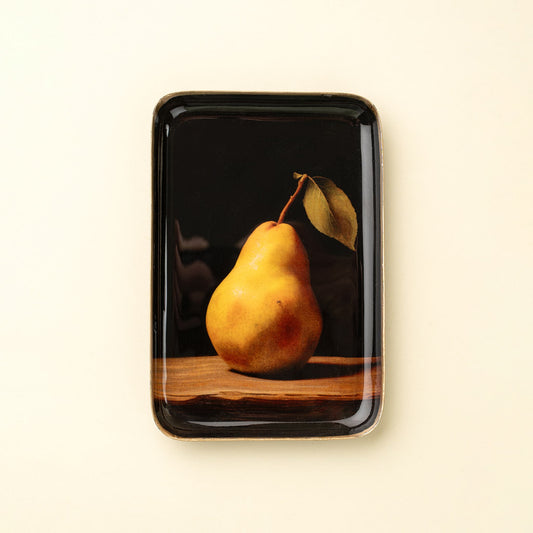 A rectangular glossy black ceramic plate featuring a classic still life print of a yellow pear with a leaf on a wooden surface, set against a warm cream background.