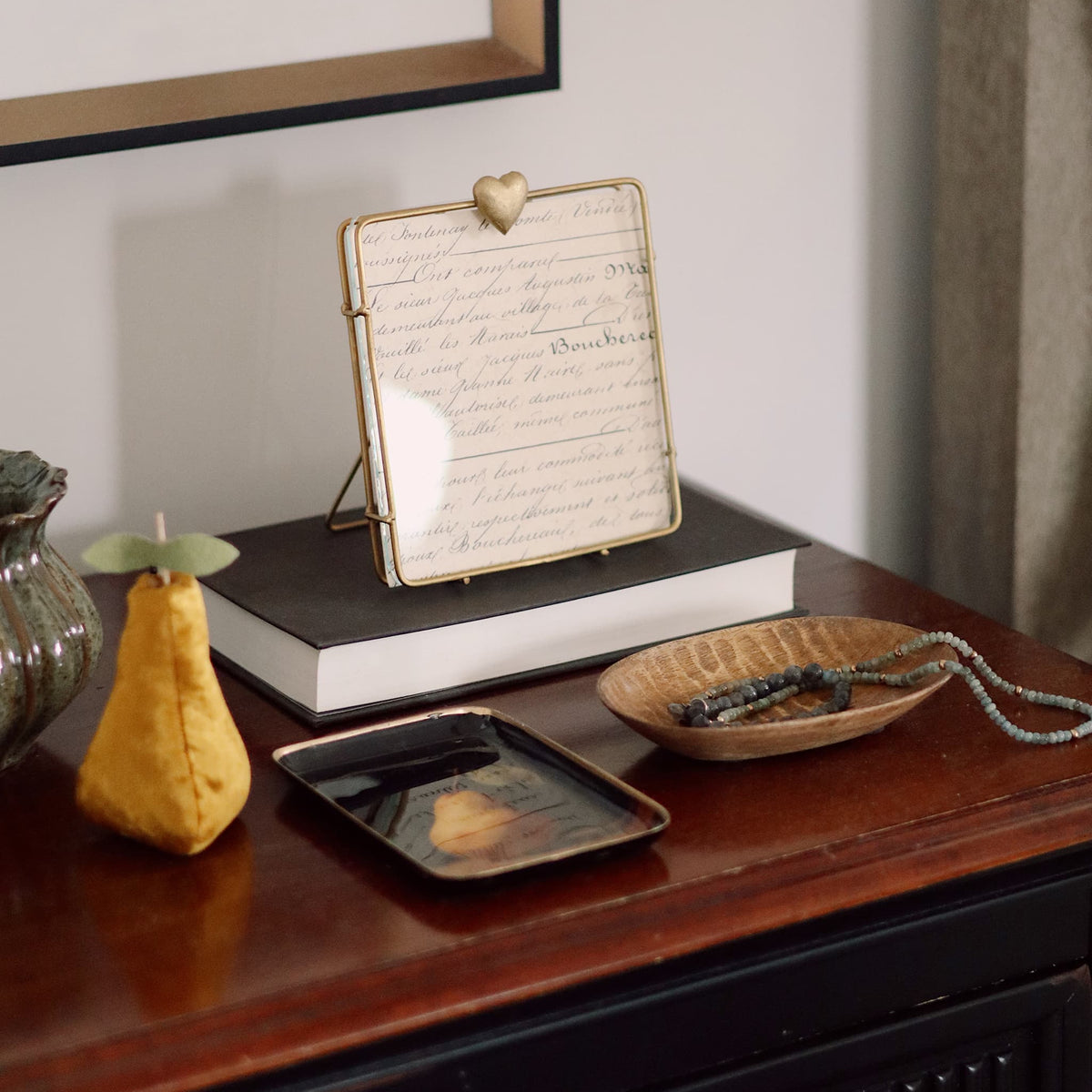 A square decorative picture frame with a gold metal border and stand, holding a vintage-style cursive script card, styled on a dark wooden chest alongside a yellow pear, a small tray, and a bowl.