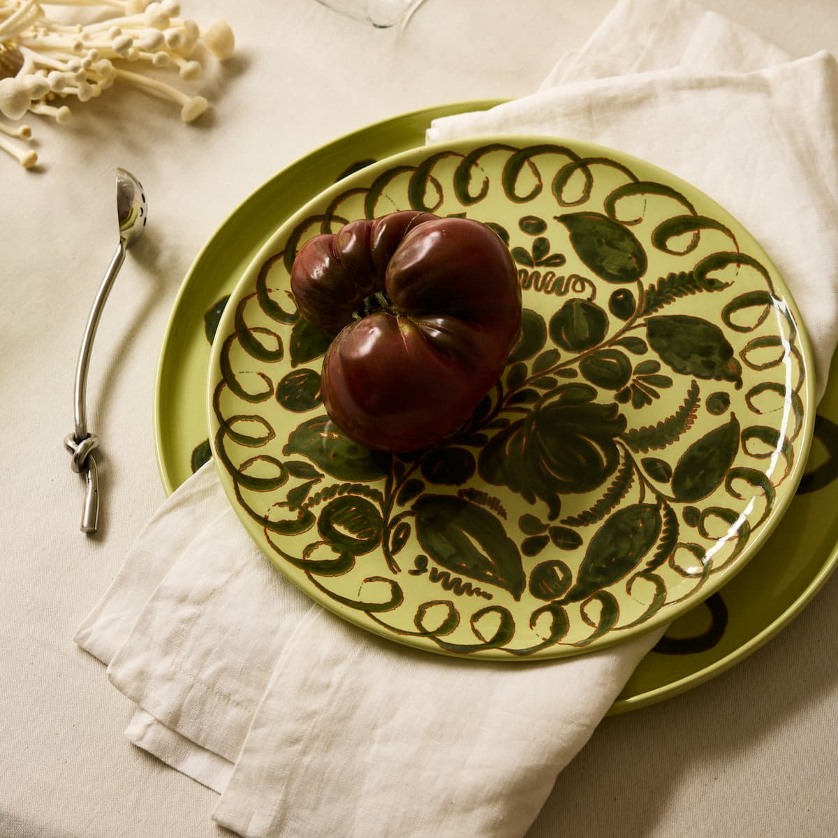 Heirloom tomato on a set of green hand-painted dishes with a table linen and a pickle spoon, laid on a white surface.