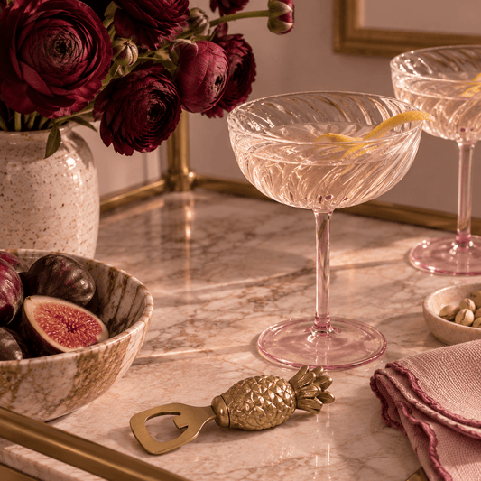 A pink champagne coupe cocktail glass with a lemon twist, set on a marble bar counter next to a gold pineapple-shaped bottle opener, a bowl of figs, and a vase of burgundy flowers.