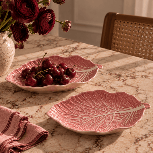 Two pink cabbage leaf ceramic serving platters on a marble table, one filled with cherries and the other empty, with a floral arrangement and woven chair in the background.