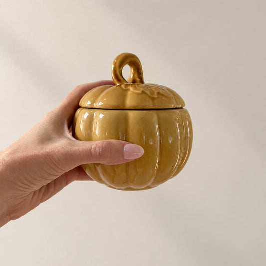 Pumpkin-shaped ceramic bowl with lid, in golden yellow ochre, held in front of a white background.