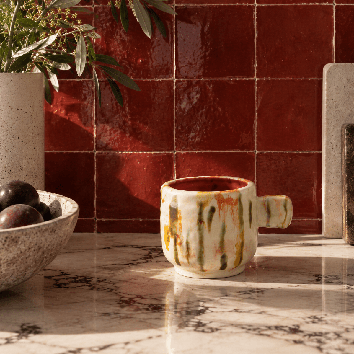 A ceramic mug with a handle, featuring a cream-colored exterior with green, orange, and yellow vertical brushstroke patterns and a deep red interior, set on a marble countertop against a red tiled wall.
