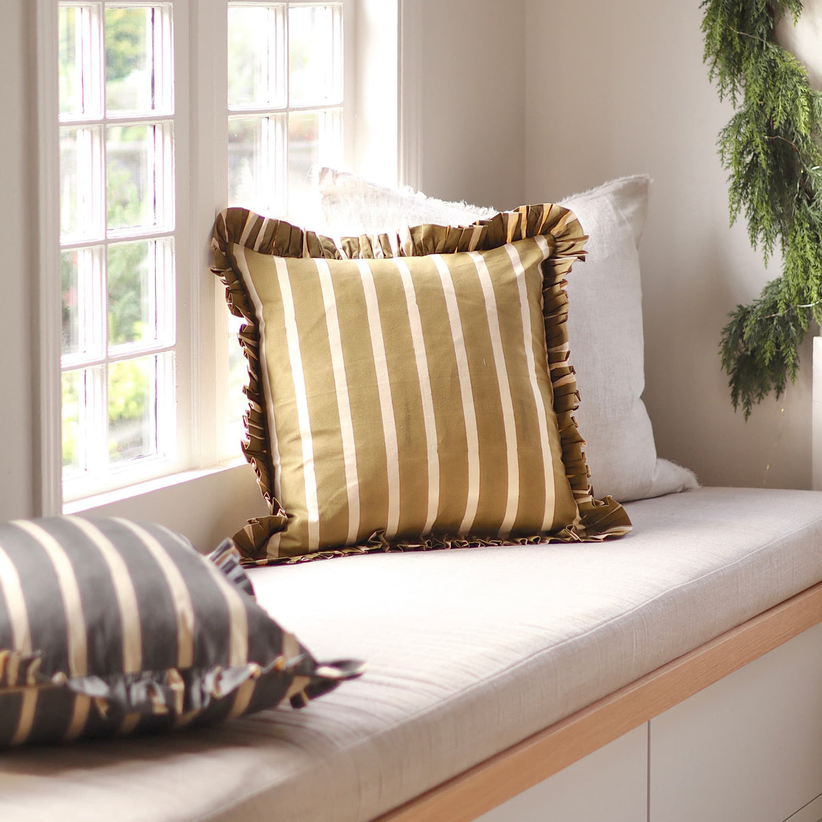 A styled interior scene showing a window seat cushion set. The main product is a square throw pillow with vertical stripes in olive green and cream, featuring a ruffled edge.
