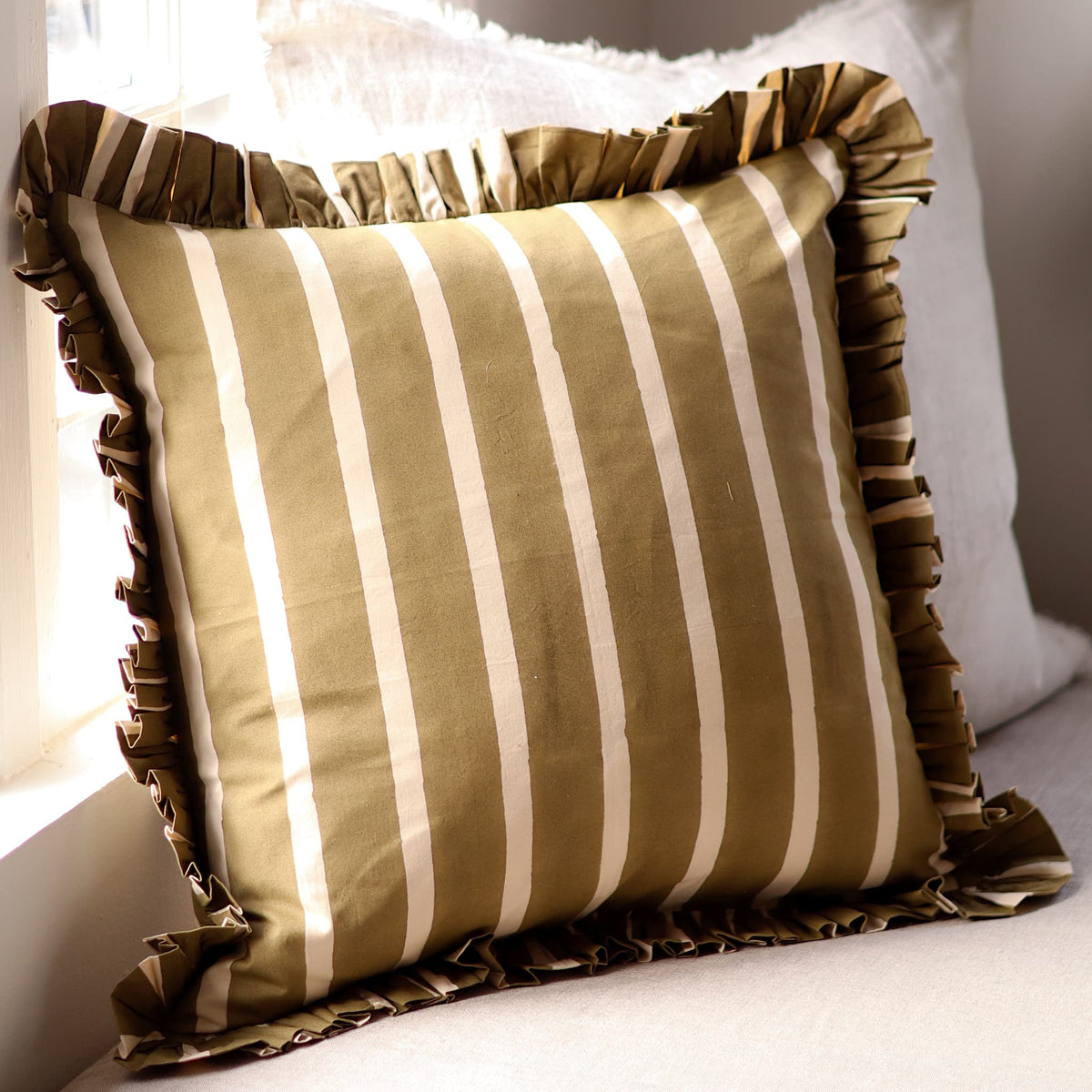 A square throw pillow with vertical olive green and cream stripes, featuring a ruffled fabric border, styled on a light-coloured cushion in front of a white pillow near a window.