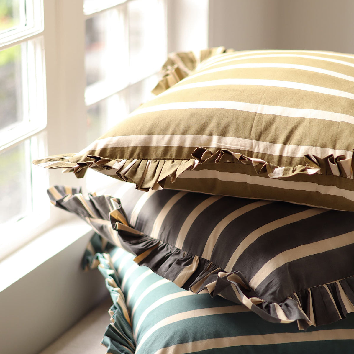 Three stacked ruffled striped pillows in olive green, charcoal gray, and teal blue, placed on a windowsill in natural light.