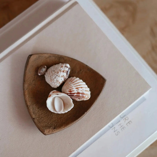 A small metal dish holding three seashells, placed on a beige card inside a white frame over a light wood surface.